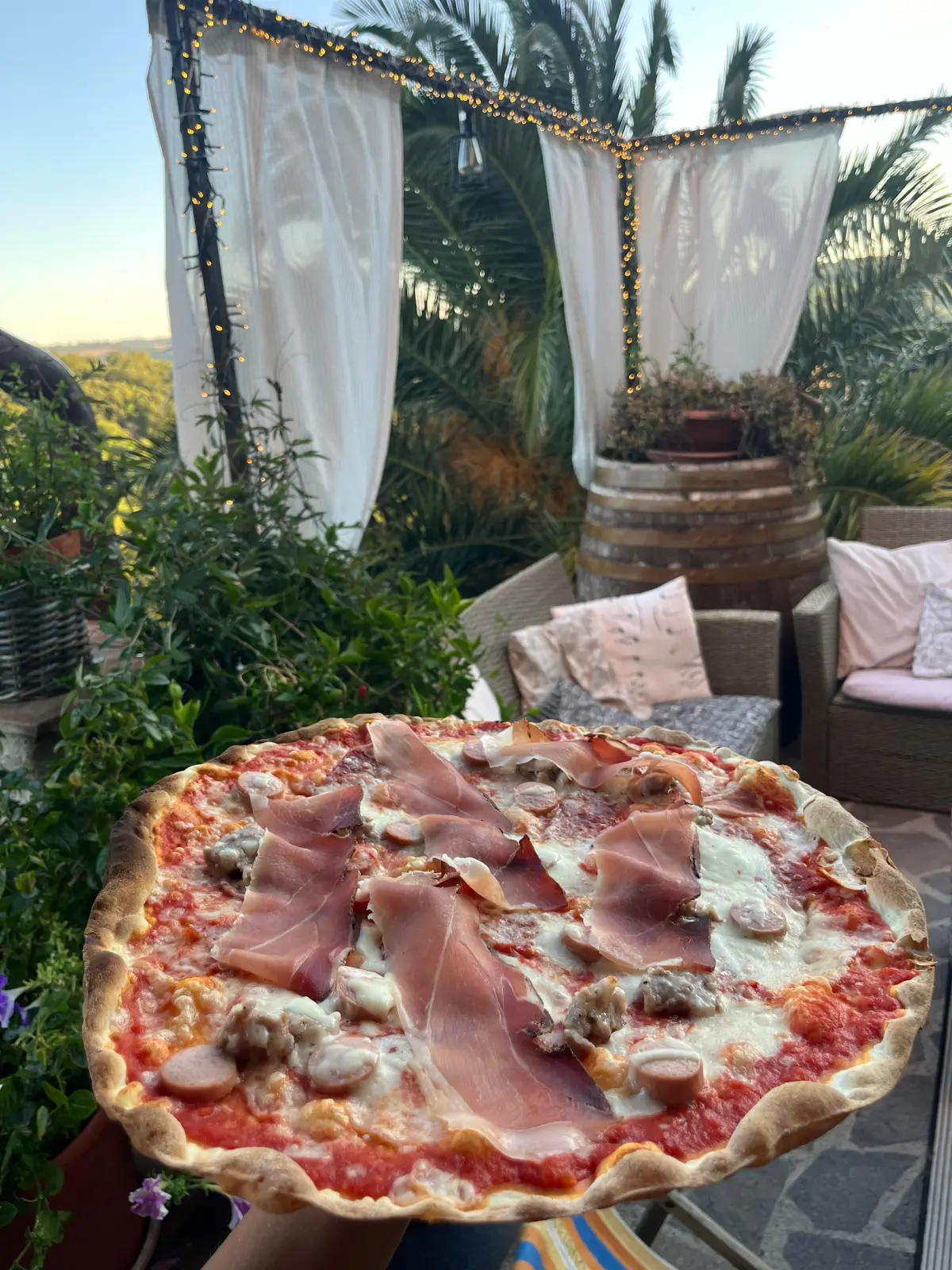 Pizza con prosciutto crudo servita in terrazza tra palme e tende bianche